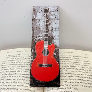 Könnte beinhalten: Lesezeichen mit roter Gitarre und Notenhintergrund. Das Lesezeichen zeigt eine leuchtend rote Gitarrenillustration vor einem Hintergrund aus Noten und einem strukturierten, neutral getönten Hintergrund. Ein tolles Geschenk.