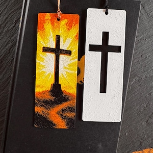 Könnte beinhalten: Zwei rechteckige Lesezeichen mit Kreuzmotiven. Eines zeigt ein Kreuz auf einem Hügel mit orange-gelbem Sonnenuntergangshintergrund. Das andere ist weiß mit einem ausgeschnittenen Kreuz. Beide haben Quasten. Die Lesezeichen liegen auf einem geschlossenen Buch.