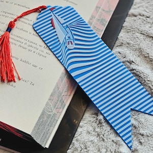 Könnte beinhalten: Blau-weiß gestreifter Lesezeichen mit Segelboot-Design. Das Lesezeichen hat eine rote Quaste. Es liegt auf einem offenen Buch, ideal für Leseratten.