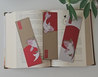 May include: Three decorative bookmarks featuring a koi fish design. The bookmarks have a red and tan color scheme with a Japanese-inspired aesthetic. The word "Koi" is printed on the bookmarks. The bookmarks are placed on an open book.