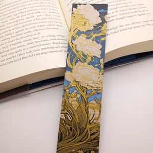 Könnte beinhalten: Ein rechteckiger Lesezeichen mit einem floralen Design aus weißen Blumen, grünem Laub und blauem Hintergrund. Das Lesezeichen befindet sich in einem offenen Buch, was auf seine Verwendung hindeutet.