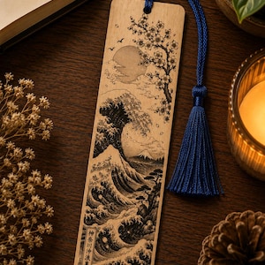 May include: A rectangular wooden bookmark featuring a detailed laser-engraved illustration of a large ocean wave, Mount Fuji, a full moon, and cherry blossom branches. The bookmark includes a blue silk tassel attached to the top with a cord.