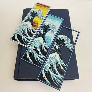 May include: Three rectangular bookmarks featuring the iconic Japanese artwork "The Great Wave off Kanagawa." Each bookmark depicts a different color scheme of the wave and a setting sun or moon. The bookmarks are resting on a dark blue book.