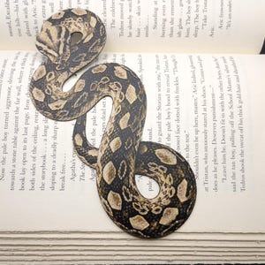 Può includere: Un segnalibro a forma di serpente con un motivo dettagliato di squame marroni, beige e nere, posto su un libro aperto. Un accessorio di lettura.