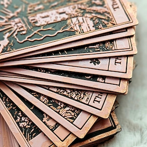 Peut inclure: Un jeu de cartes de tarot avec une finition cuivrée. Les cartes présentent des motifs détaillés avec des accents noirs et cuivrés. Le texte "The Tower" est visible sur une carte. Les cartes sont étalées, mettant en valeur l'œuvre d'art.