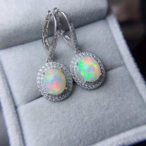 May include: A pair of silver-toned hoop earrings with oval opal gemstones. Each opal is surrounded by a halo of small, clear crystals. The opals display a play of color, with hues of pink, green, and yellow.