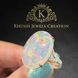 May include: An oval opal ring with a silver band and diamond detailing. The opal displays a milky white hue with colourful specks of orange, pink, and green. The ring is set against a black backdrop with the text "Khushi Jewels Creation".