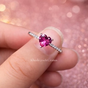 May include: A silver ring featuring a heart-shaped, vibrant pink gemstone. The band is adorned with small, clear stones. The text "Khushi jewels Creation" is visible on the image.