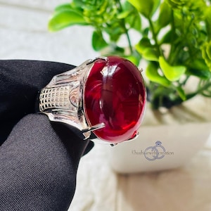 May include: A silver ring with a large, red, oval-shaped gemstone. The ring is set in a silver band with a textured design. The ring is on a black surface.