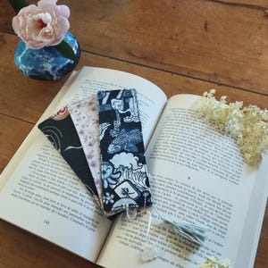 Peut inclure: Quatre marque-pages en tissu à motifs japonais reposent sur un livre ouvert. Les marque-pages présentent des motifs de carpes koï, de fleurs de cerisier et de formes géométriques en bleu, noir et blanc. Un vase avec une fleur rose est en arrière-plan.