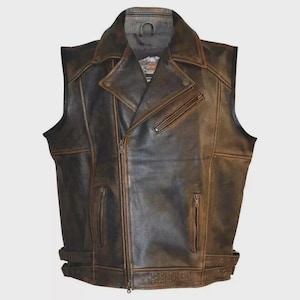 Może przedstawiać: Brązowa skórzana kamizelka w klasycznym stylu motocyklowym. Kamizelka posiada kołnierz z klapami, asymetryczne zapięcie na zamek błyskawiczny i wiele kieszeni zapinanych na zamek. Logo Harley-Davidson jest wytłoczone na dole. Kamizelka ma postarzany wygląd.