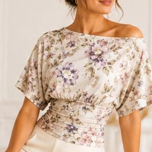 May include: A cream-colored blouse featuring a floral pattern in shades of purple, pink, and green. The top has an off-the-shoulder neckline, short sleeves, and a wide, gathered, fitted waistband. It is paired with solid cream-colored trousers.