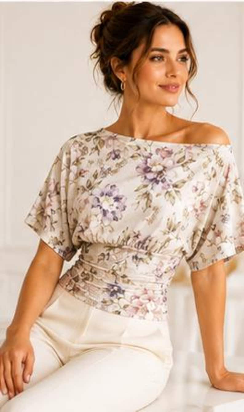 May include: A cream-colored blouse featuring a floral pattern in shades of purple, pink, and green. The top has an off-the-shoulder neckline, short sleeves, and a wide, gathered, fitted waistband. It is paired with solid cream-colored trousers.