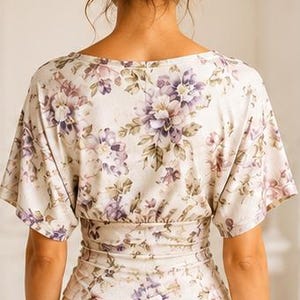 May include: A short-sleeved top featuring a cream base with a purple and brown floral pattern. The garment has a wide neckline, loose sleeves, and a fitted, gathered waist section that creates a cinched silhouette above plain cream trousers.