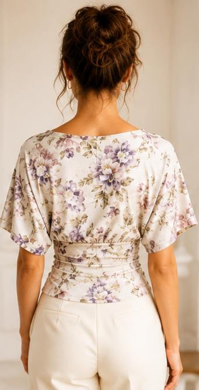May include: A short-sleeved top featuring a cream base with a purple and brown floral pattern. The garment has a wide neckline, loose sleeves, and a fitted, gathered waist section that creates a cinched silhouette above plain cream trousers.