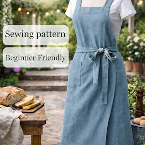 Peut inclure: Un tablier en lin bleu clair avec un design enveloppant, noué à la taille. Le tablier a une poche et est présenté sur un mannequin. Le texte sur l'image indique "Natural Linen Apron", "Sewing pattern" et "Beginner Friendly".