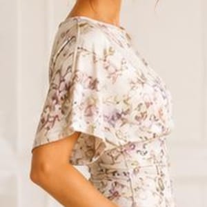 May include: A short-sleeved blouse featuring a cream base with a delicate floral pattern in shades of purple and green, paired with solid cream-colored trousers. The blouse has a relaxed fit with wide sleeves and a fitted waistline detail.