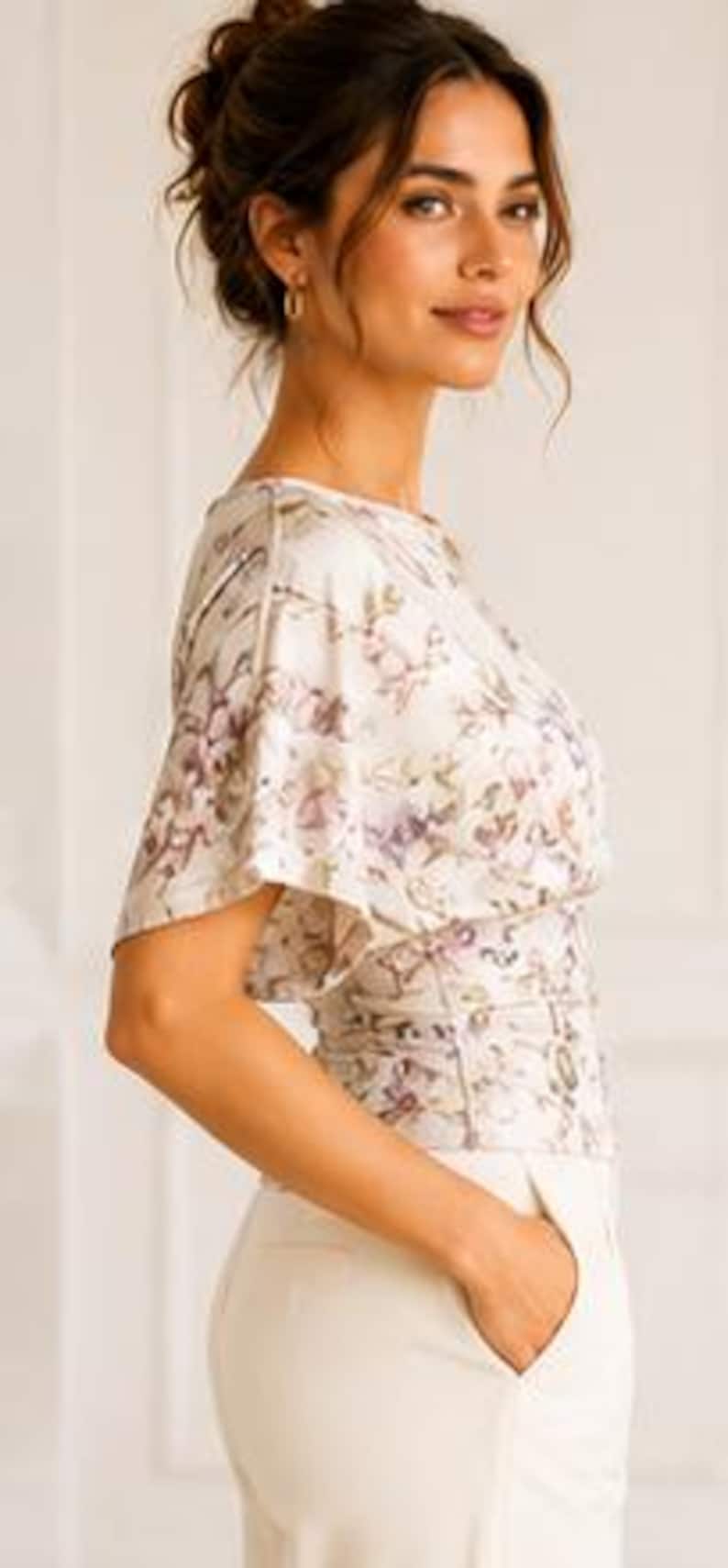 May include: A short-sleeved blouse featuring a cream base with a delicate floral pattern in shades of purple and green, paired with solid cream-colored trousers. The blouse has a relaxed fit with wide sleeves and a fitted waistline detail.