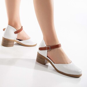 May include: White leather Mary Jane style shoes with a brown ankle strap and a wooden block heel. The shoes have a rounded toe and a buckle closure. The heel height is approximately 2.5 inches.