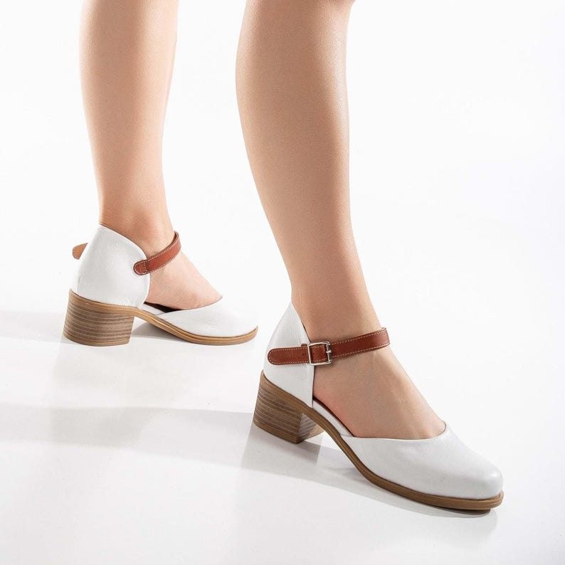 May include: White leather Mary Jane style shoes with a brown ankle strap and a wooden block heel. The shoes have a rounded toe and a buckle closure. The heel height is approximately 2.5 inches.