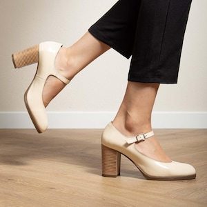 May include: Cream-colored Mary Jane heels with a square toe and a chunky, wooden-look heel. The shoes have a strap that buckles across the top of the foot. The heels appear to be approximately 3 inches high.