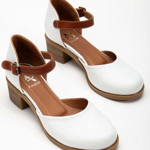 May include: A pair of white leather Mary Jane style shoes with brown ankle straps and block heels. The shoes have a rounded toe and the brand name "Analin" is visible on the insole.