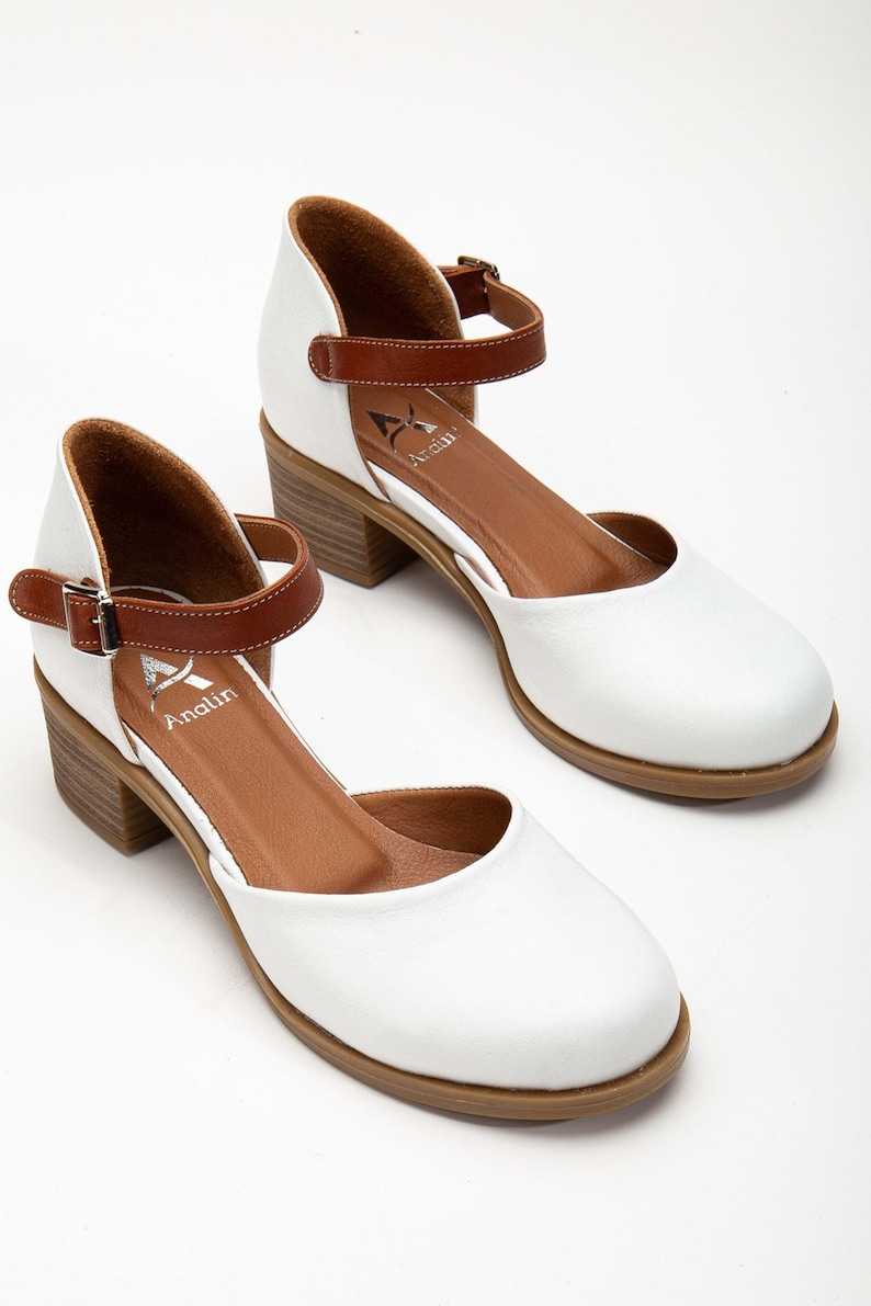 May include: A pair of white leather Mary Jane style shoes with brown ankle straps and block heels. The shoes have a rounded toe and the brand name "Analin" is visible on the insole.