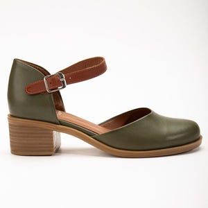 May include: Olive green leather Mary Jane style shoe with a brown leather ankle strap and a small silver buckle. The shoe has a block heel and a rounded toe. The sole is a light brown color.