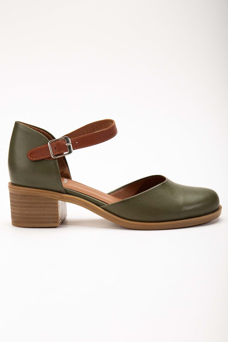 May include: Olive green leather Mary Jane style shoe with a brown leather ankle strap and a small silver buckle. The shoe has a block heel and a rounded toe. The sole is a light brown color.