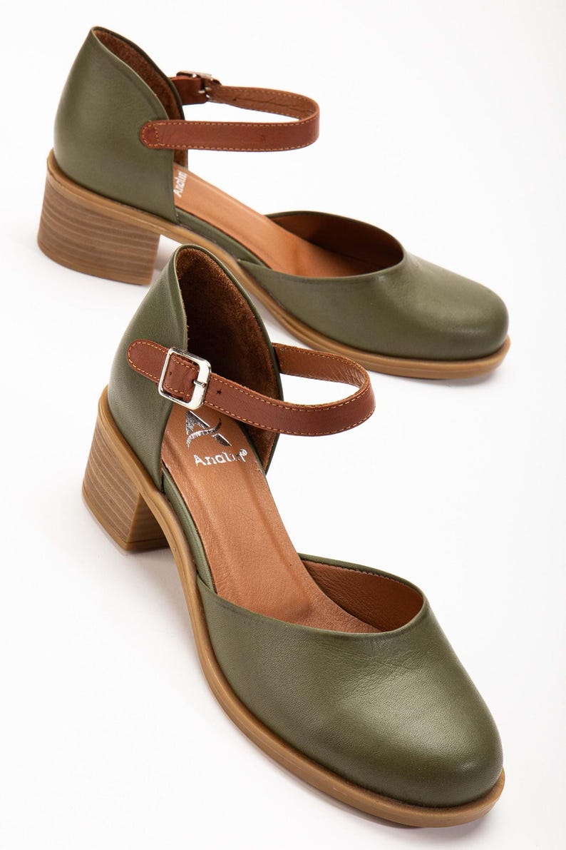 May include: Olive green leather Mary Jane style shoes with a brown ankle strap and a wooden block heel. The shoes have a rounded toe and a buckle closure. The brand name "Anali" is visible on the insole.