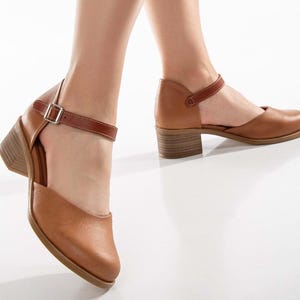 May include: A pair of brown leather shoes with a block heel and an ankle strap. The shoes have a closed toe design and a buckle closure on the strap. The heels are approximately 2 inches high.