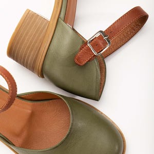 May include: Olive green leather sandals with a brown leather ankle strap and a wooden block heel. The sandals have a rounded toe and a silver buckle. The interior is a light brown color.