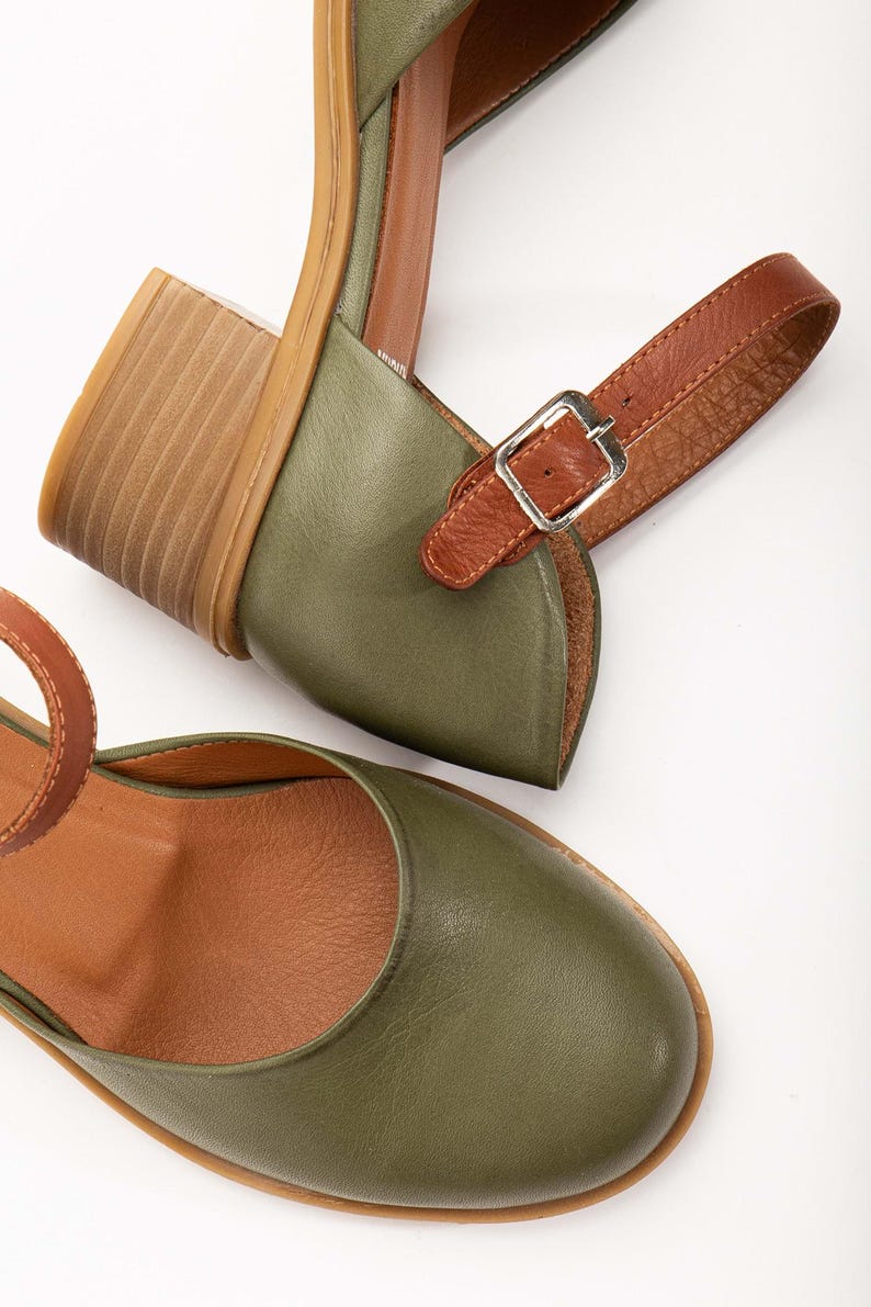 May include: Olive green leather sandals with a brown leather ankle strap and a wooden block heel. The sandals have a rounded toe and a silver buckle. The interior is a light brown color.