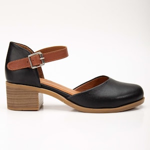 May include: Black leather Mary Jane style shoe with a brown ankle strap and buckle closure. The shoe has a rounded toe and a wooden block heel. The sole is a light brown color.