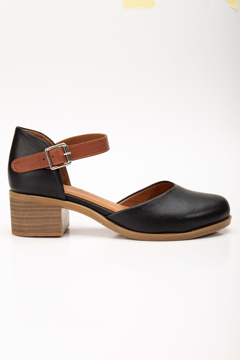 May include: Black leather Mary Jane style shoe with a brown ankle strap and buckle closure. The shoe has a rounded toe and a wooden block heel. The sole is a light brown color.