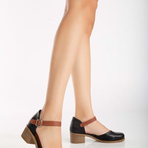 May include: Black leather Mary Jane style shoes with a brown ankle strap and a small wooden heel. The shoes have a rounded toe and a buckle closure. The shoes are worn by a person.
