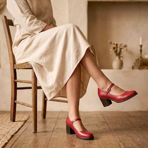 May include: A pair of red Mary Jane style shoes with a block heel. The shoes have a strap that buckles across the top of the foot. The shoes are made of a smooth material and have a dark brown heel. The shoes are on a wooden floor.