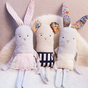 May include: Three handmade plush bunny toys. One bunny is wearing a pink lace skirt, another has a black and white striped shirt, and the third bunny is plain. The bunnies are sitting on a white fluffy surface.
