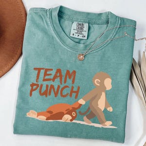 May include: A teal Comfort Colors t-shirt with the words "TEAM PUNCH" in orange. The shirt features a cartoon monkey pulling a brown animal. A silver necklace with a pendant is draped across the shirt.