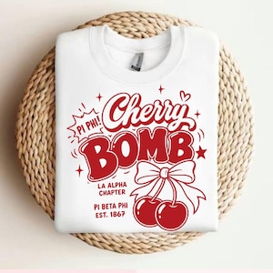 May include: White crew neck sweatshirt with a red graphic that says "Cherry Bomb" with cherries and a bow. The text "Pi Phi!" and "LA Alpha Chapter Pi Beta Phi Est. 1867" are also in red. The sweatshirt is on a woven surface.