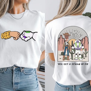 May include: A white t-shirt featuring a front graphic of a fist bump between a yellow-patterned arm and a white robotic arm. The back graphic shows a desert scene with a cowboy and a space ranger, with the text YOU GOT A FRIEND IN ME.