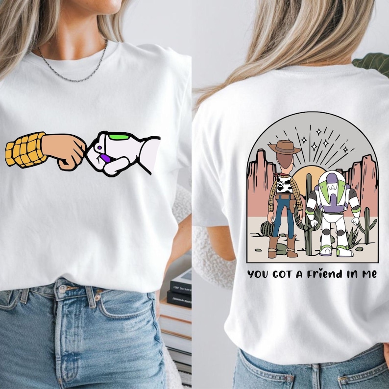 May include: A white t-shirt featuring a front graphic of a fist bump between a yellow-patterned arm and a white robotic arm. The back graphic shows a desert scene with a cowboy and a space ranger, with the text YOU GOT A FRIEND IN ME.