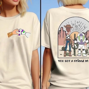 May include: A cream-colored t-shirt featuring a small graphic on the front of two hands forming a heart shape. The back displays a desert scene with two cartoon characters, cacti, and the text "YOU GOT A FRIEND IN ME" printed in black lettering.
