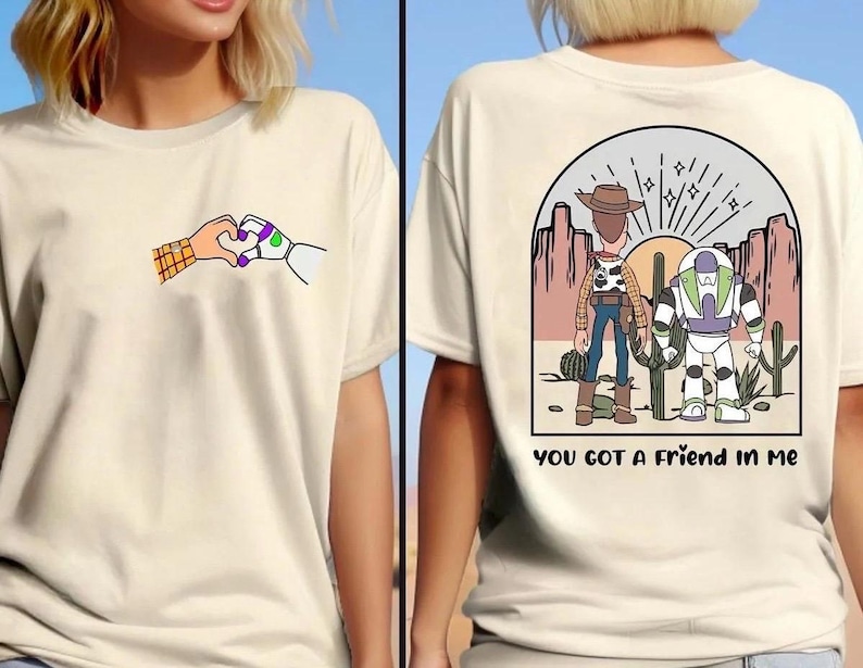 May include: A cream-colored t-shirt featuring a small graphic on the front of two hands forming a heart shape. The back displays a desert scene with two cartoon characters, cacti, and the text "YOU GOT A FRIEND IN ME" printed in black lettering.