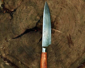 May include: A Damascus steel chef's knife with a wooden handle. The blade has a unique pattern, and the handle is a warm, reddish-brown color. The knife is resting on a weathered, circular wooden surface.