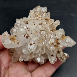 May include: A cluster of clear quartz crystals with brown inclusions. The crystals are tightly packed together, forming a textured, irregular shape. The quartz is held in a hand against a dark background.