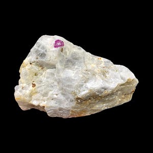 Op de afbeelding: Een ruw, gebroken wit mineraal specimen met een kleine, levendige roze edelsteen ingebed in het oppervlak. De steen heeft een kristallijne structuur en een licht verweerde uitstraling, tegen een zwarte achtergrond.