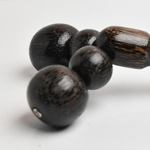 May include: A dark brown wooden massage tool with a series of connected spheres and a handle. The spheres vary in size, and the handle has a pointed end. The wood grain is visible, and the tool appears to be smooth and polished.