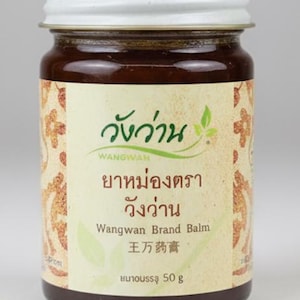May include: A glass jar of Wangwan Brand Balm with a white lid. The jar has a label with text in multiple languages, including Thai and English. The balm inside is a dark brown color, and the jar contains 50 g of product.