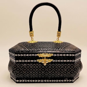 May include: A black woven handbag with a curved black handle and gold-colored accents. The rectangular bag features a decorative clasp and intricate woven patterns in black and white. The handle is attached with ornate gold-colored hardware.